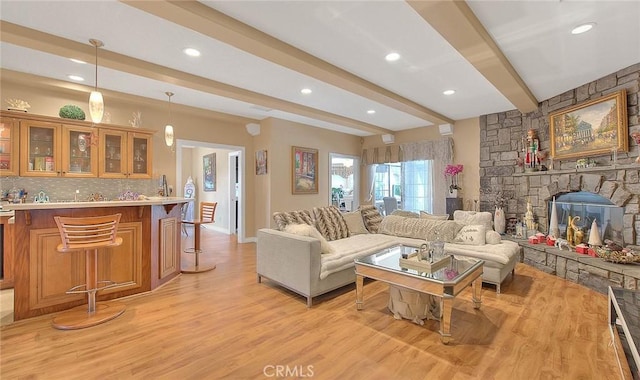 living room featuring beam ceiling, light wood-style flooring, recessed lighting, a stone fireplace, and wet bar