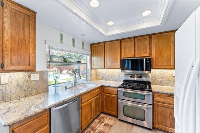 kitchen with brown cabinets, ornamental molding, a sink, a tray ceiling, and appliances with stainless steel finishes