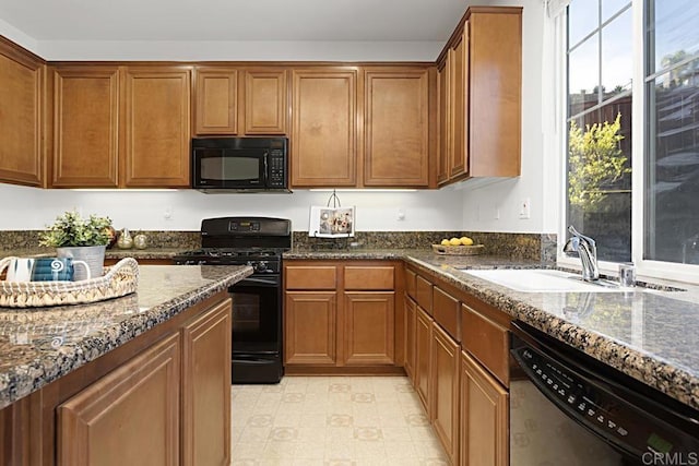 kitchen featuring light floors, dark stone counters, brown cabinets, black appliances, and a sink