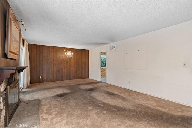 unfurnished living room featuring visible vents, wooden walls, a stone fireplace, carpet flooring, and an inviting chandelier