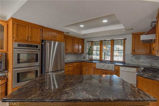 kitchen with brown cabinetry, a tray ceiling, a sink, appliances with stainless steel finishes, and tasteful backsplash