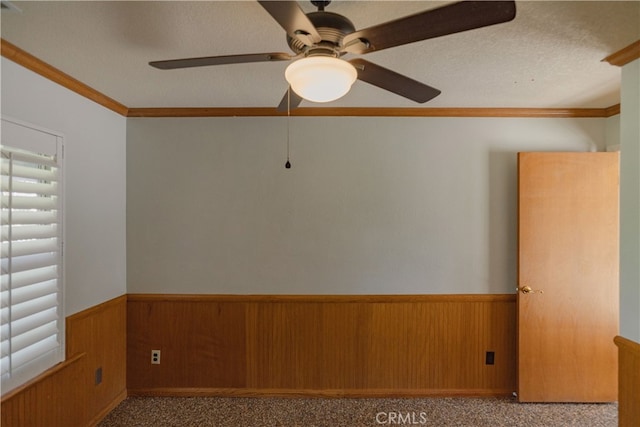 carpeted spare room with crown molding, a wainscoted wall, wood walls, a textured ceiling, and a ceiling fan