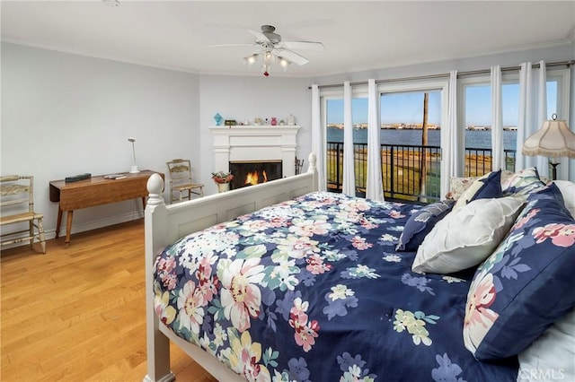 bedroom with access to exterior, a water view, ornamental molding, a warm lit fireplace, and wood finished floors