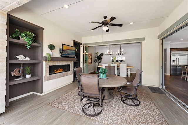 dining area with baseboards, built in features, ceiling fan with notable chandelier, wood finished floors, and a glass covered fireplace