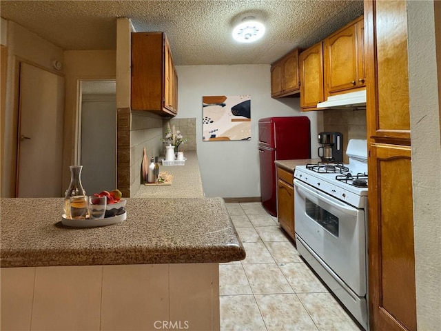 kitchen with under cabinet range hood, gas range gas stove, light tile patterned flooring, brown cabinetry, and decorative backsplash