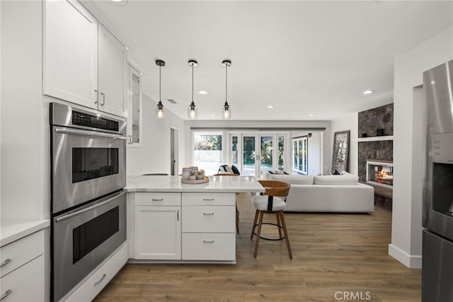 kitchen with stainless steel double oven, a peninsula, light countertops, white cabinets, and a large fireplace