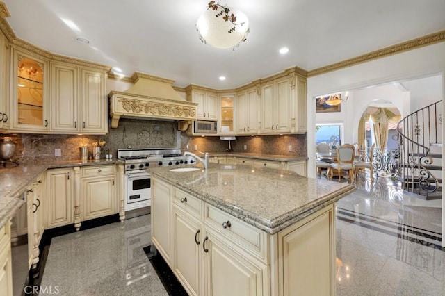 kitchen featuring cream cabinetry, custom range hood, granite finish floor, arched walkways, and appliances with stainless steel finishes