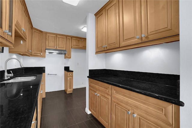 kitchen featuring dark tile patterned floors, a sink, under cabinet range hood, dark stone counters, and brown cabinetry