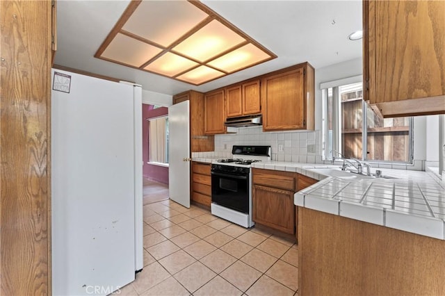 kitchen with tasteful backsplash, tile counters, under cabinet range hood, gas range, and freestanding refrigerator