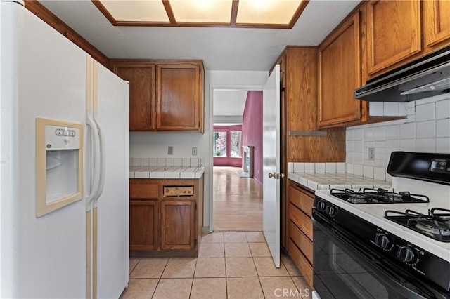 kitchen featuring under cabinet range hood, tile countertops, white fridge with ice dispenser, light tile patterned floors, and gas range