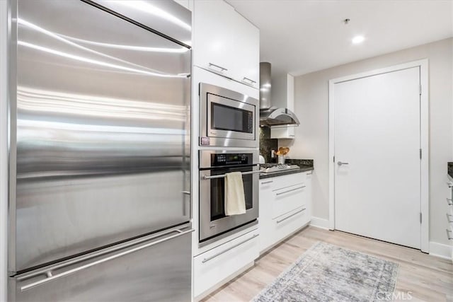 kitchen with light wood-style flooring, built in appliances, wall chimney exhaust hood, and white cabinets