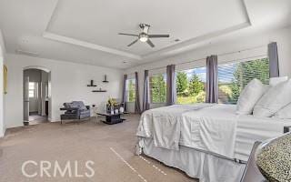 carpeted bedroom featuring a tray ceiling, arched walkways, multiple windows, and ceiling fan