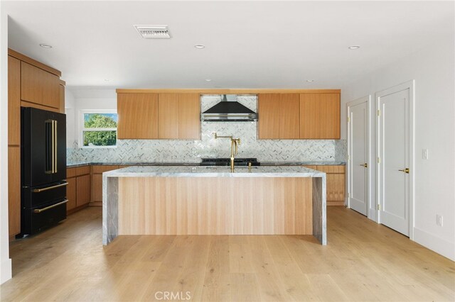 kitchen with light wood-type flooring, an island with sink, tasteful backsplash, high quality fridge, and wall chimney range hood