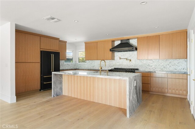 kitchen featuring wall chimney range hood, an island with sink, light wood-style floors, modern cabinets, and high end black fridge