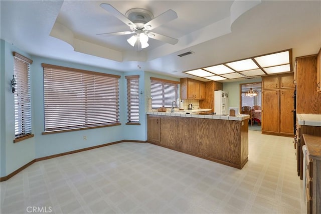 kitchen with white fridge with ice dispenser, a peninsula, brown cabinetry, a raised ceiling, and tile counters