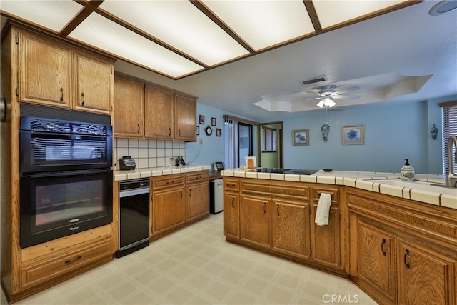 kitchen with brown cabinets, black appliances, a tray ceiling, tasteful backsplash, and tile counters