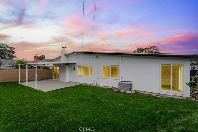 back of house at dusk with fence, cooling unit, a chimney, a yard, and a patio area