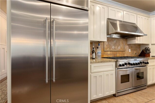 kitchen featuring light tile patterned floors, high end appliances, light stone countertops, under cabinet range hood, and backsplash