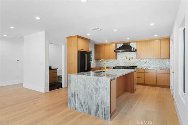 kitchen featuring visible vents, light stone counters, wall chimney exhaust hood, light wood finished floors, and high end black fridge