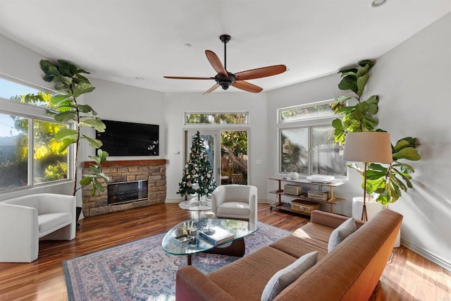 living room featuring ceiling fan, baseboards, a stone fireplace, and wood finished floors