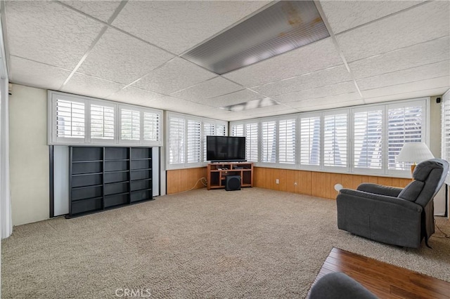 living area with a drop ceiling, plenty of natural light, carpet floors, and wood walls