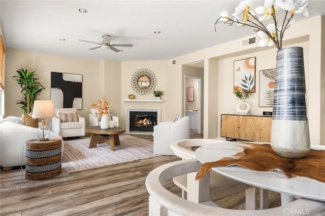 living room with a glass covered fireplace, visible vents, wood finished floors, and ceiling fan