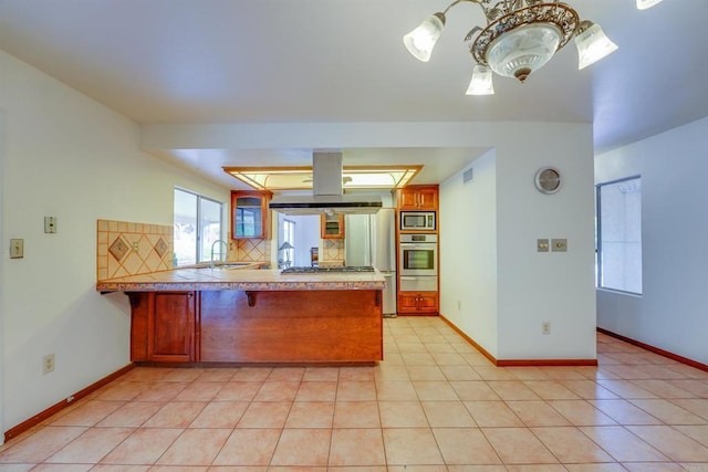 kitchen with light tile patterned floors, a peninsula, appliances with stainless steel finishes, backsplash, and island range hood