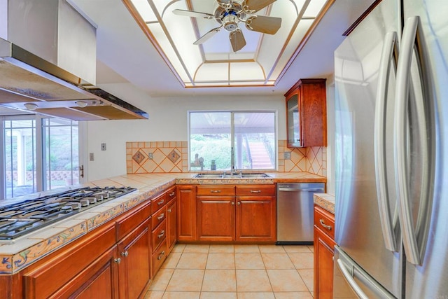 kitchen with a sink, a tray ceiling, appliances with stainless steel finishes, light tile patterned flooring, and wall chimney range hood