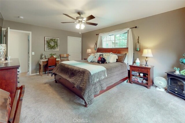 bedroom with a wood stove, light colored carpet, and ceiling fan