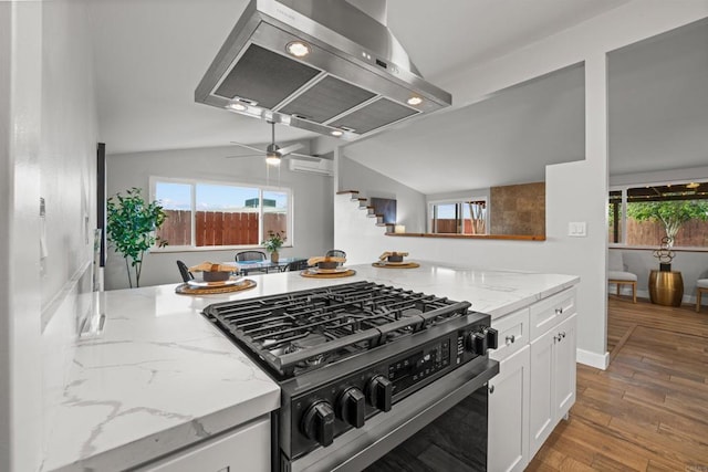 kitchen with wood finished floors, vaulted ceiling, white cabinets, gas range, and wall chimney range hood