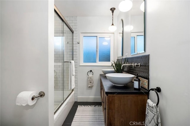 full bath with a sink, backsplash, shower / bath combination with glass door, double vanity, and baseboards