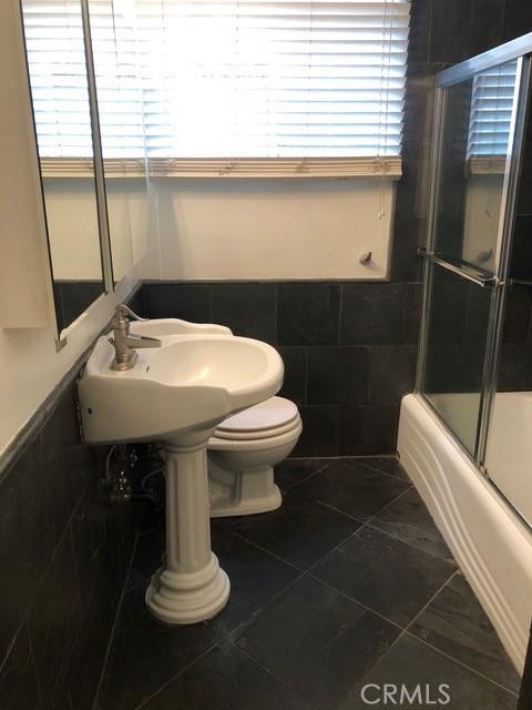 full bathroom featuring a wainscoted wall, tile patterned flooring, tile walls, toilet, and combined bath / shower with glass door