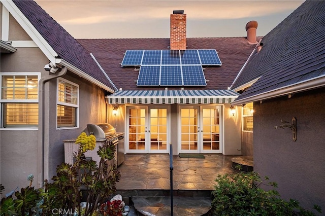 back of house at dusk with roof with shingles, roof mounted solar panels, stucco siding, french doors, and a patio