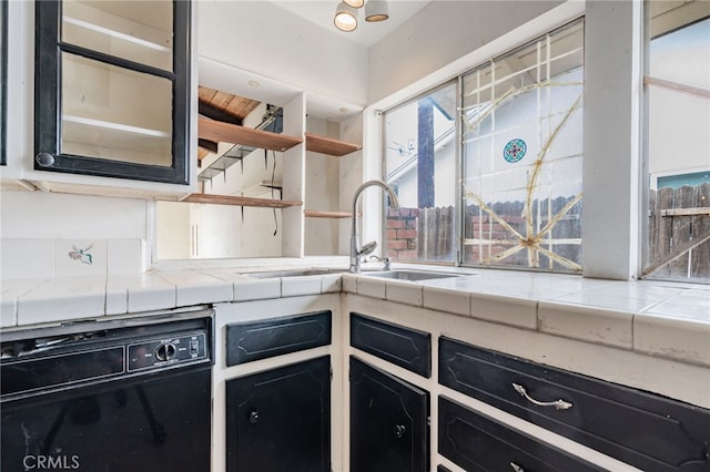 kitchen with dark cabinetry, tile countertops, open shelves, a sink, and black dishwasher