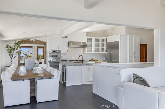 kitchen with stainless steel appliances, white cabinets, wall chimney range hood, glass insert cabinets, and vaulted ceiling with beams