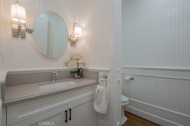 half bathroom featuring vanity, toilet, wood finished floors, and a wainscoted wall