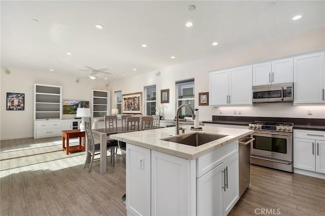 kitchen with wood finished floors, a center island with sink, a sink, appliances with stainless steel finishes, and white cabinetry