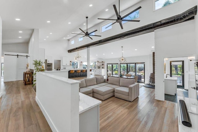 living room with recessed lighting, ceiling fan, light wood-style floors, a towering ceiling, and a barn door