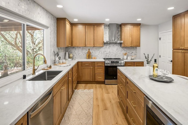 kitchen featuring brown cabinetry, light wood finished floors, a sink, stainless steel appliances, and wall chimney range hood