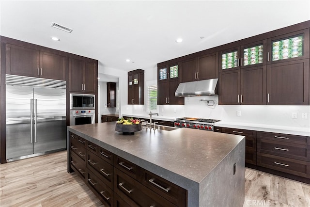 kitchen with under cabinet range hood, dark countertops, built in appliances, and a sink