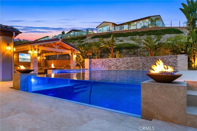 pool at dusk with fence, a gazebo, a fire pit, a fenced in pool, and a patio area