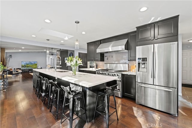 kitchen with light stone countertops, a center island with sink, stainless steel appliances, wall chimney exhaust hood, and dark cabinets