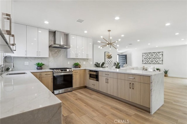 kitchen with a peninsula, a sink, stainless steel gas range oven, wall chimney range hood, and modern cabinets