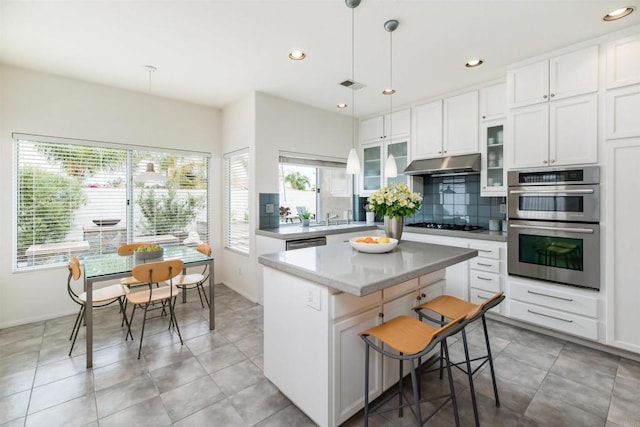 kitchen featuring under cabinet range hood, tasteful backsplash, a kitchen island, appliances with stainless steel finishes, and glass insert cabinets