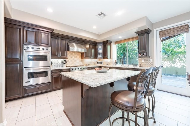 kitchen with a kitchen bar, visible vents, ventilation hood, stainless steel appliances, and dark brown cabinets