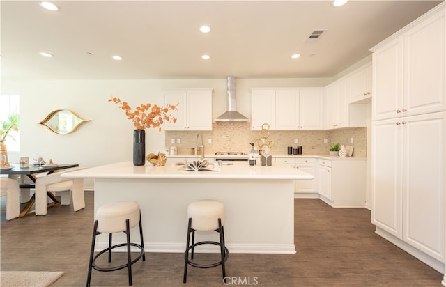 kitchen with visible vents, wall chimney range hood, an island with sink, dark wood-style floors, and white cabinetry