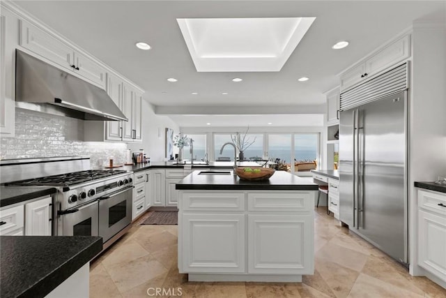 kitchen featuring high end appliances, a sink, white cabinets, under cabinet range hood, and dark countertops