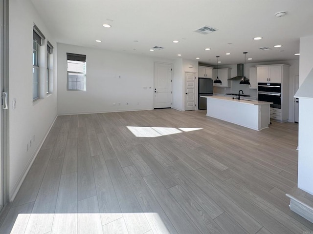 kitchen featuring a center island with sink, light wood-type flooring, open floor plan, and wall chimney range hood