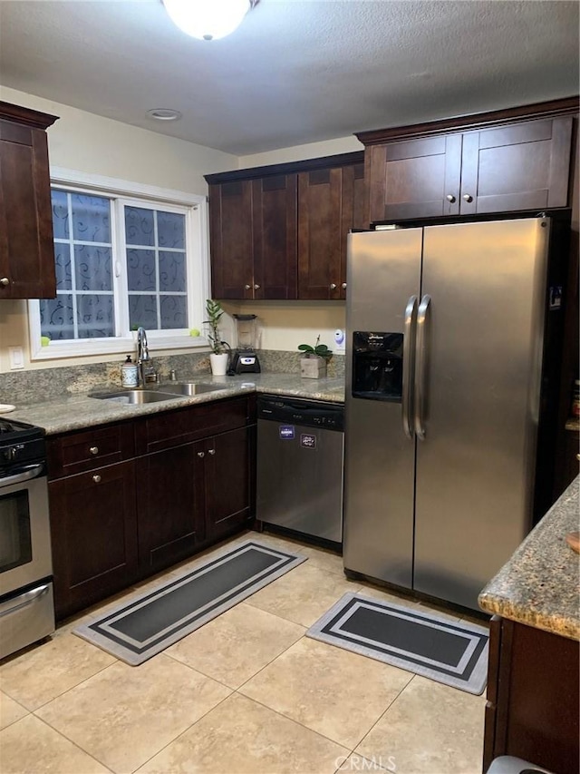 kitchen with stone countertops, a sink, appliances with stainless steel finishes, light tile patterned floors, and dark brown cabinets