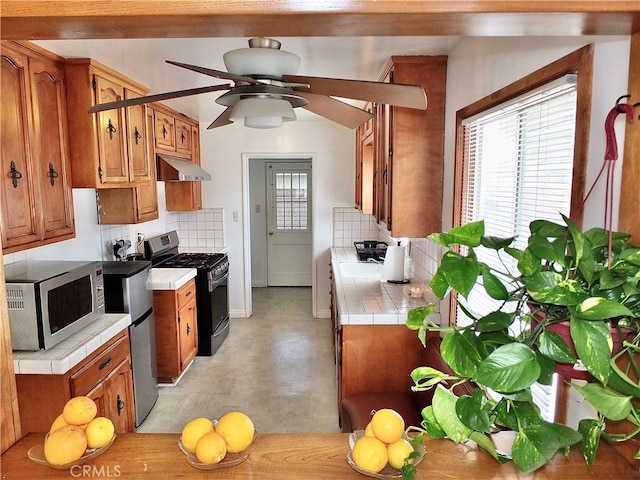 kitchen with brown cabinets, under cabinet range hood, tasteful backsplash, stainless steel appliances, and tile counters